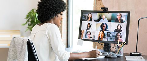 A woman works at a desk and takes notes while on a video call with nine other people