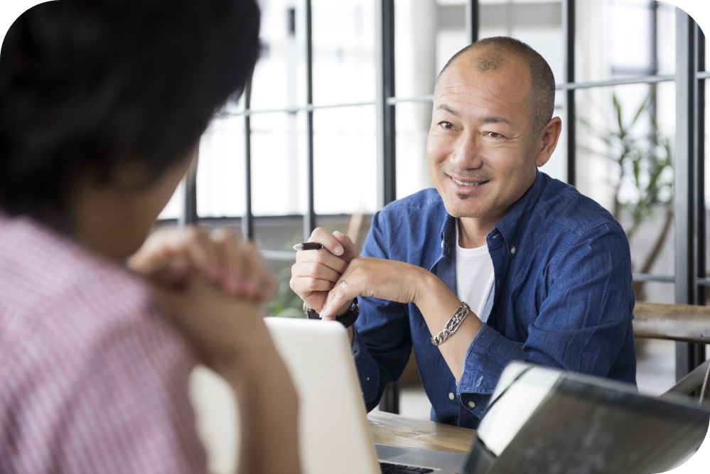 Man in a blue shirt sitting at a desk while talking to another person sitting across from them