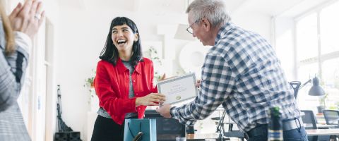 A professional in a bright red shirt, laughing, hands another professional in a plaid button-up an award certificate over their desk