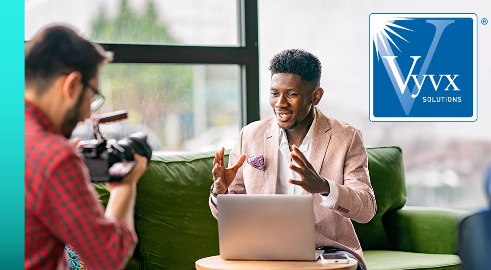  Two people sitting across from each other with a laptop open in front of them with one person gesturing and speaking while the other person holds a camera 