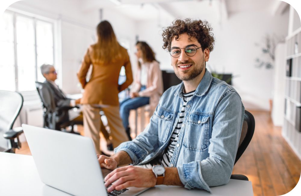 Person in casual attire sitting in an office setting smiling and using a laptop.