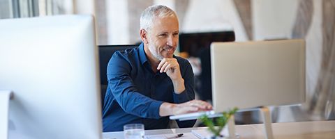 Person in a dark blue button up shirt sitting at a desk smiling and using a laptop.