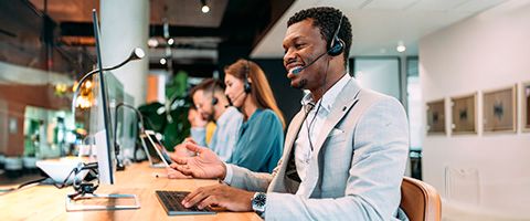 Two people wearing headsets work at computers in an office while a third person talks on a mobile phone 