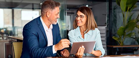 A smiling business professional engages in a discussion with a colleague using a laptop with windows in the background