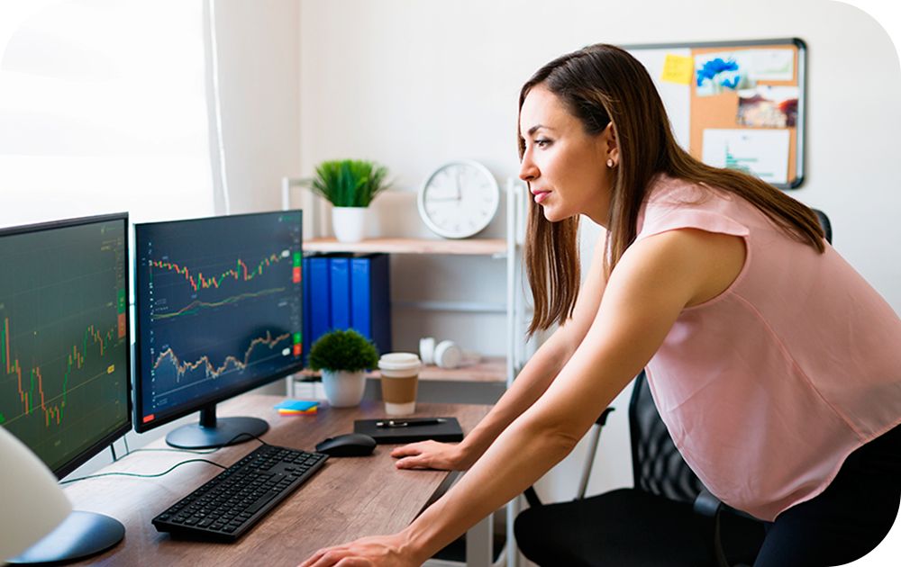 An individual in a home office leans over a desk and looks at financial charts displayed on two monitors