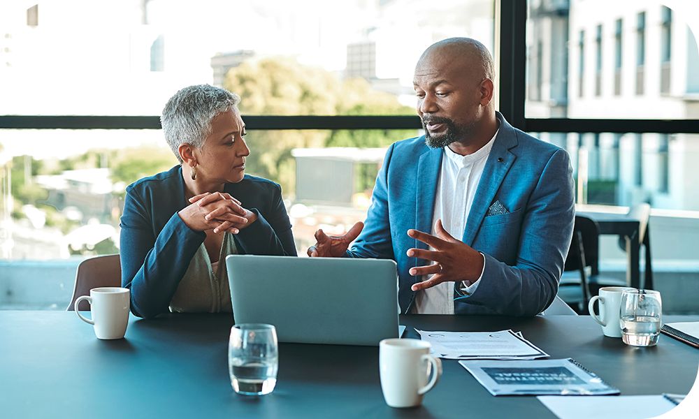 Two professionals sit at a table with an open laptop in front of them as they converse.