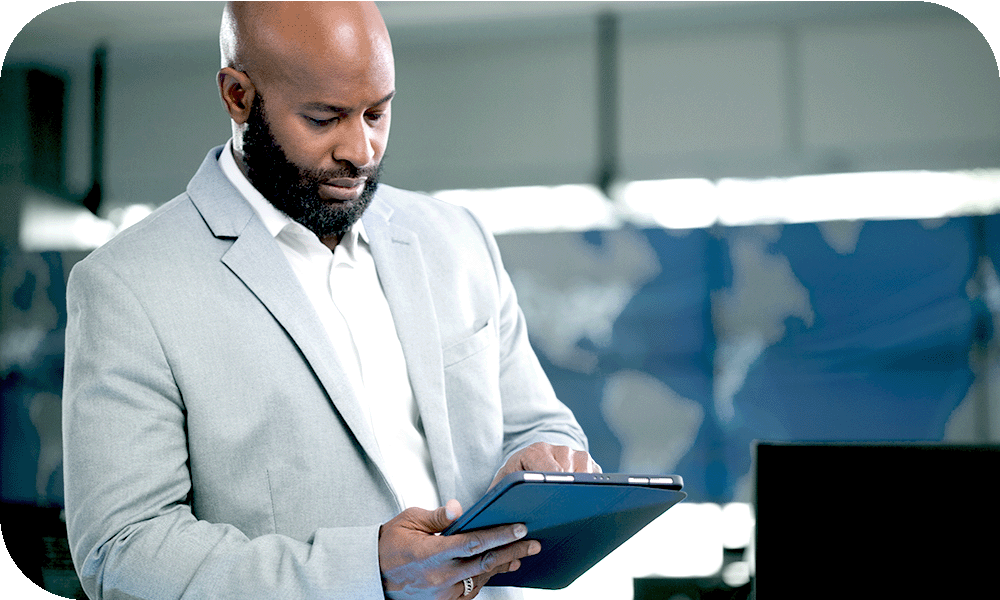  Man with tablet in hand standing in front of large global maps examining information on the screen. 