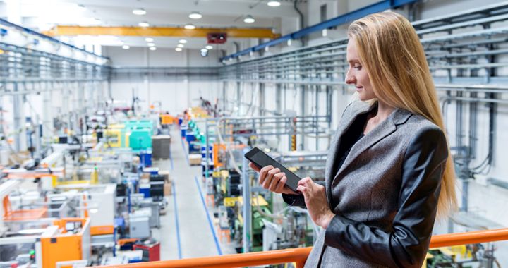 Business woman standing above a factory floor in a warehouse while working on a tablet
