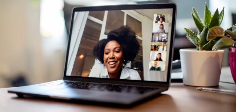 A laptop screen shows a woman utilizing the Zoom video conference feature to speak with six other people in remote locations.