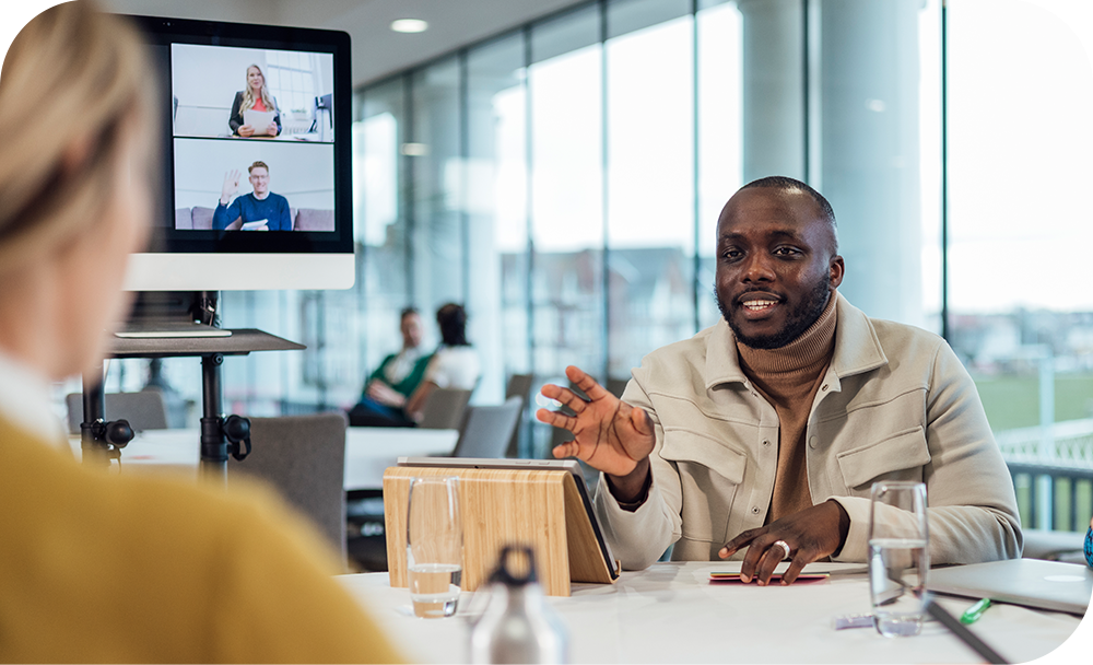 Man sitting at conference table discussing ideas with colleagues with Zoom chat on monitors in background.